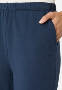 Námořnické modré tepláky vyrobené z měkké látky; mají elastický pás, boční kapsy a vyšitý logo "LES LUNES" na boku.