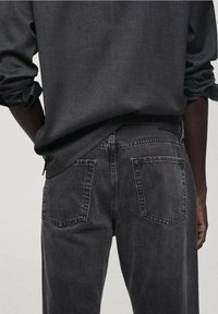 Jeans in denim nero con taglio dritto, dotati di due tasche posteriori e cuciture a contrasto. Indossati con una camicia scura, girati di spalle.