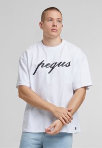 Fehér pamut póló, amelyen a "pegus" fekete írásos logó található elöl, rövid ujjakkal és laza szabással. Világoskék farmert visel.