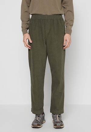 Persona che indossa pantaloni verde oliva con vita elasticizzata, maglia marrone a maniche lunghe e scarpe da ginnastica grigie, in piedi davanti a uno sfondo chiaro uniforme.