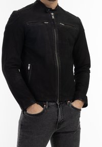 Mann in einer schwarzen Lederjacke mit Reißverschluss und Reißverschluss-Taschen auf der Brust und an den Seiten, die Hände in den Taschen, kombiniert mit dunkelgrauen Jeans.