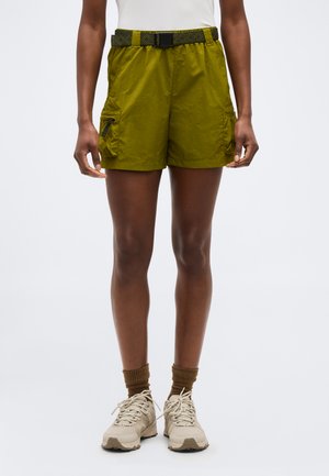 Személy olíva zöld cargo rövidnadrágban fekete övvel, bézs sportcipőben és barna zokniban, deréktól lefelé fehér háttér előtt ábrázolva.