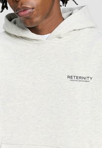 Ljusgrå hoodie av mjukt material, med en kenguruficka, dragsko i huvan och broderad text "RETERNITY CREATIVE DEPARTMENT."