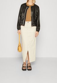 Sort faux læder bomberjakke med hvid kant, brun top, cremefarvet maxiskørt med slidse foran, gul håndtaske og sorte platform sandaler.