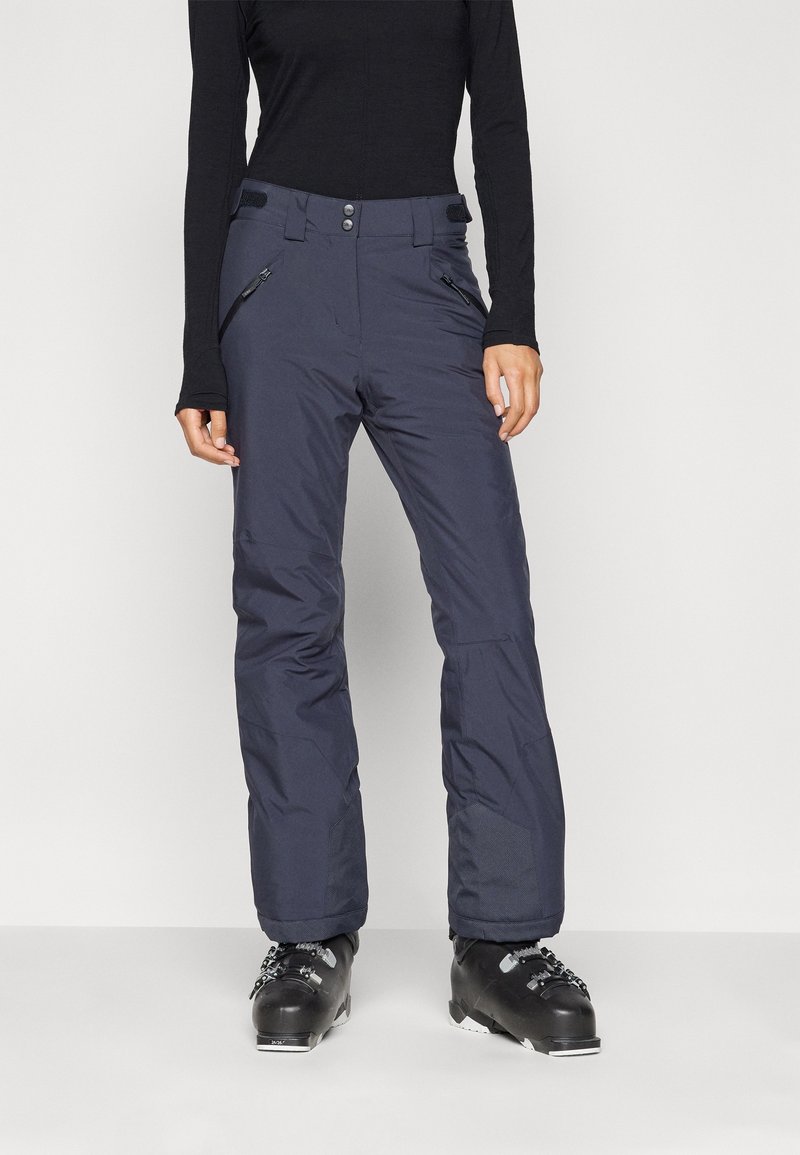 Persona con pantalones de esquí azul marino y botas de esquí negras, de pie contra un fondo claro y liso.