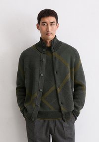 Knit cardigan in dark green with a textured pattern, featuring a high collar, button closure, and yellow diagonal stripes. Worn over a matching sweater.