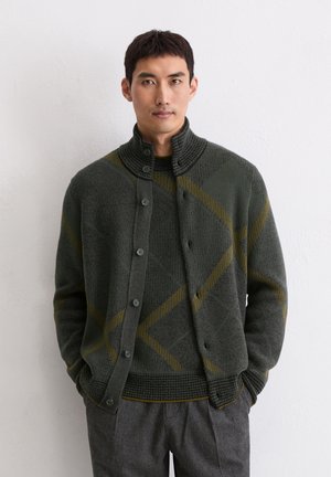 Gebreide cardigan in donkergroen met een gestructureerd patroon, voorzien van een hoge kraag, knoopsluiting en gele diagonale strepen. Draagt over een bijpassende trui.