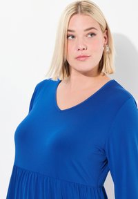 Niebieska sukienka w serek z gładkiego materiału, z plisowaną spódnicą. Krótkie rękawy, luźny krój i subtelny połysk materiału.