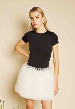 Tânără cu păr blond lung, purtând cercei în formă de inimă, bluză neagră cu mâneci scurte și fustă albă pufoasă din tull, pozând cu mâna pe șold.