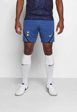 Blaue Sportshorts mit einem strukturierten Muster, ausgestattet mit gelben Nike- und Team-Logos. Getragen mit weißen bis zu den Knien reichenden Socken und weißen Sneakers.