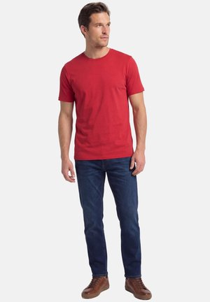 Mann trägt ein schlichtes rotes Kurzarm-T-Shirt, dunkelblaue Jeans und braune Schnürschuhe, schaut nach links.