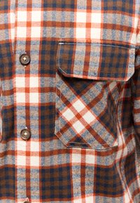 Kariertes Flanellhemd mit orangefarbenen, braunen und blauen Karomustern, mit Knopfleiste und einer Brusttasche links. Die Textur ist weich.