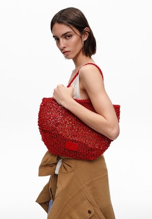 Joven mujer con el pelo corto y marrón sosteniendo un gran bolso rojo de punto para el hombro, vestida con chaqueta beige atada a la cintura y top blanco sin mangas.