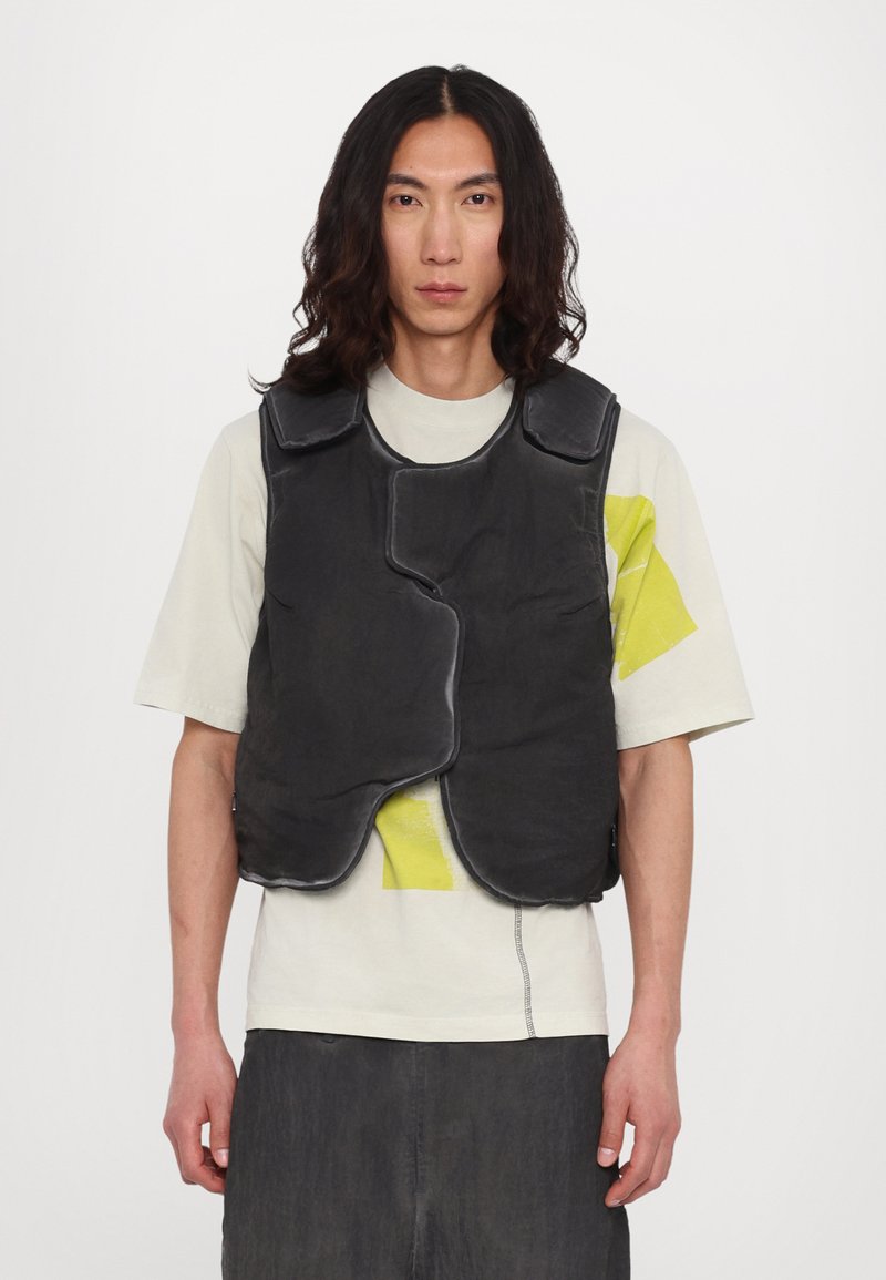 Gilet rembourré noir avec épaules arrondies, doté d'une fermeture éclair et de deux poches latérales. Porté par-dessus un t-shirt de couleur claire.