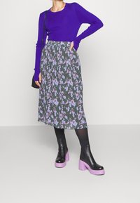 Lila ribbad långärmad topp, blommig grön midi kjol, svarta ankelskor med lila klackar. Modellen poserar med en hand på höften.