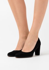 Escarpins en daim noir à talons hauts, avec un bout rond et un talon épais, présentant une texture lisse et un design minimaliste.