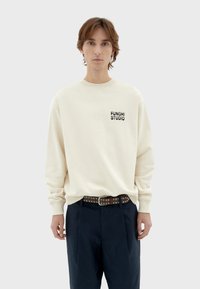 Crème sweatshirt met ribgebreide manchetten en hals, voorzien van een zwart "FUNGHISTUDIO" logo op de borst, gecombineerd met donkere broek en een gesp met studs.