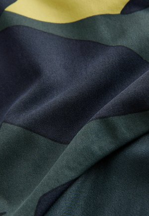 Textil med överlappande geometriska former i mörkgrön, marinblå och gul. Slät, vävd textur med en lätt lyster.