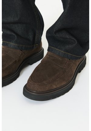 Mocassins en daim marron avec des semelles noires épaisses portés avec un jean bleu foncé, coutures visibles sur le jean et les chaussures, sur fond blanc.