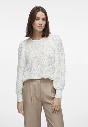 Blusa bianca con texture e motivo ondulato, scollatura tonda e maniche lunghe a palloncino, abbinata a pantaloni beige a vita alta.