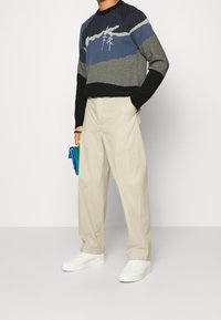 Personne portant un pull à plusieurs tons de bleu et noir avec un motif d'arbre, pantalon large beige, baskets blanches, tenant une petite pochette bleue et verte.