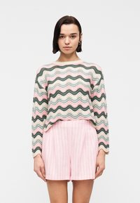 Γυναίκα με κοντά σκούρα μαλλιά που φορεί κρίκους στα αυτιά, ένα πουλόβερ με παστέλ ριγέ κυματιστή σχεδίαση και ροζ ριγέ σορτς με ψηλή μέση, στέκεται κοιτώντας προς τα εμπρός.