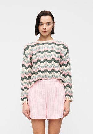 Woman with short dark hair wearing hoop earrings, a pastel wavy striped sweater, and pink striped high-waist shorts, standing facing forward.