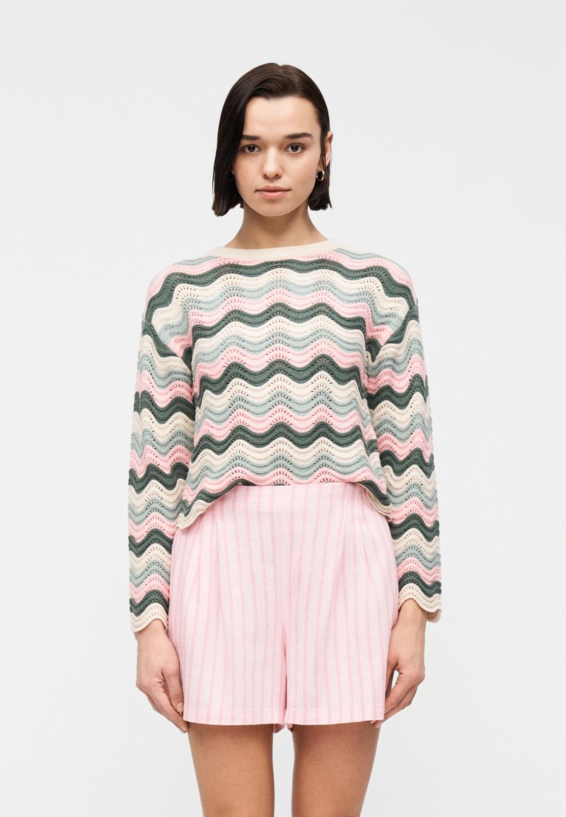 Γυναίκα με κοντά σκούρα μαλλιά που φορεί κρίκους στα αυτιά, ένα πουλόβερ με παστέλ ριγέ κυματιστή σχεδίαση και ροζ ριγέ σορτς με ψηλή μέση, στέκεται κοιτώντας προς τα εμπρός.