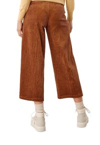 Pantalons en velours côtelé marron à jambe large avec des détails de côtes verticales. Présentent une taille haute et deux poches arrière. Portés avec des baskets de couleur claire.