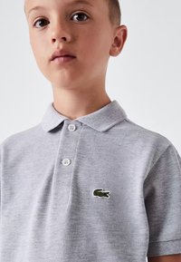 Grå pikétröja med en strukturerad väv, två knappar vid kragen och en grön broderad krokodil-logotyp på bröstet.