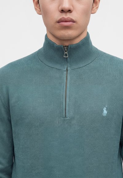 Gros plan d'une personne portant un pull zippé texturé bleu sarcelle avec un petit logo brodé sur la poitrine gauche.