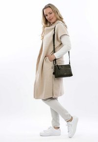 Poncho beige e morbido in tessuto, con tasche laterali, indossato sopra una maglietta a maniche lunghe bianca e pantaloni grigio chiaro, abbinato a sneakers bianche e una borsa verde scuro.