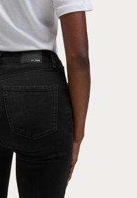 Svarta denimjeans med medelhög midja, med bakfickor och en läderetikett med "EST. 1884" på midjebandet. Slät textur.