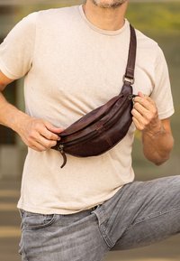 Riñonera de cuero marrón con dos cremalleras, diseño curvado y correa ajustable, llevada cruzada sobre una camisa beige.