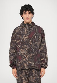 Giacca con cappuccio di un borgogna scuro con un motivo astratto e testurizzato in nero e ocra. Presenta polsini elastici e una chiusura con zip frontale.