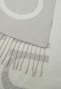 Textil de culoare gri deschis cu modele circulare și geometrice, având o franjură țesută cu ciucuri răsuciți pe o margine.