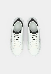 Paire de baskets basses blanches et noires avec lacets, logo sur la languette et le côté, vues de dessus sur un fond uni.