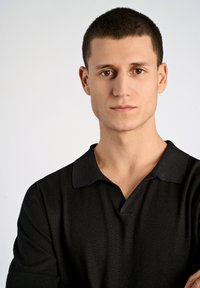 Svart polo t-shirt med ribbad textur, med djup V-hals och korta ärmar, bärs av en person med kort, mörkt hår mot en neutral bakgrund.