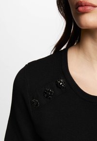 Zwarte gebreide trui met een ronde halslijn, een textuurstof en vier decoratieve zwarte bloemenknopen op de schouder.