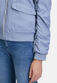Hellblaues Bomberjacke aus glattem Material mit elastischen Bündchen, mit einem Frontreißverschluss und einer großen aufgesetzten Tasche auf der linken Seite.