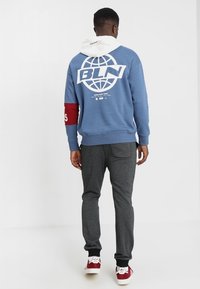 Blå sweatshirt med vit huva och stor vit globgrafik. Har en röd ärmdekoration och grå byxor med sidoficka.