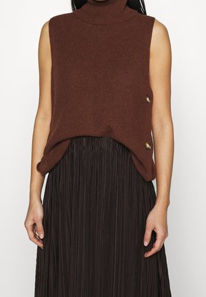 Haut sans manches marron en maille à col roulé avec deux boutons sur le côté, porté avec une jupe plissée foncée sur un mannequin féminin.