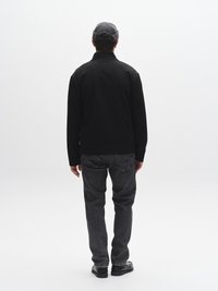 Juoda striukė su apykakle, dera su pilkomis džinsais. Asmuo dėvi pilką kepurę ir juodus batus. Fonas paprastas, baltas.