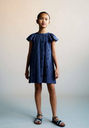 Navyblaues Baumwollkleid mit kurzen Rüschenärmeln, verziert mit bestickten Blumenmustern. Kombiniert mit blauen Sandalen mit weißen Akzenten.