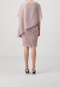 Femme portant une robe en dentelle rose pâle avec un superposé transparent asymétrique, associée à des sandales à talons hauts argentées, se tenant sur un fond uni.