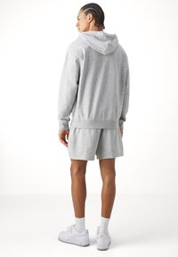 Vyras su pintomis plaukais, vilkintis pilką kapučiną, atitinkančius šortus, baltas kojines ir baltus Nike Air sportbačius, stovi atgręžęs nugarą baltame fone.