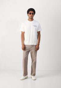 Witte katoenen t-shirt met zwarte tekst, lichtbeige broek en witte sneakers. Model draagt donkere zonnebril en staat tegen een neutrale achtergrond.