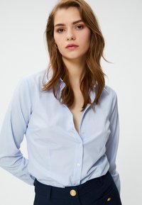 Camicia azzurra a righe con colletto classico, maniche lunghe e design aderente, abbinata a pantaloni scuri con dettagli in bottone dorato.
