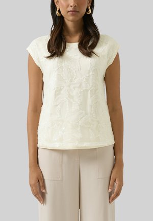 Donna che indossa una maglietta a maniche corte color panna con ricami floreali e pantaloni beige a vita alta, accessoriata con orecchini a cerchio dorati.