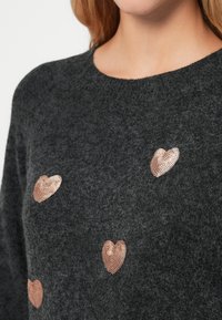 Ciemnoszara sweter dziergany z teksturowanego materiału, ozdobiony błyszczącymi, perforowanymi akcentami w kształcie serca w kolorze różowego złota, rozmieszczonymi na przodzie.